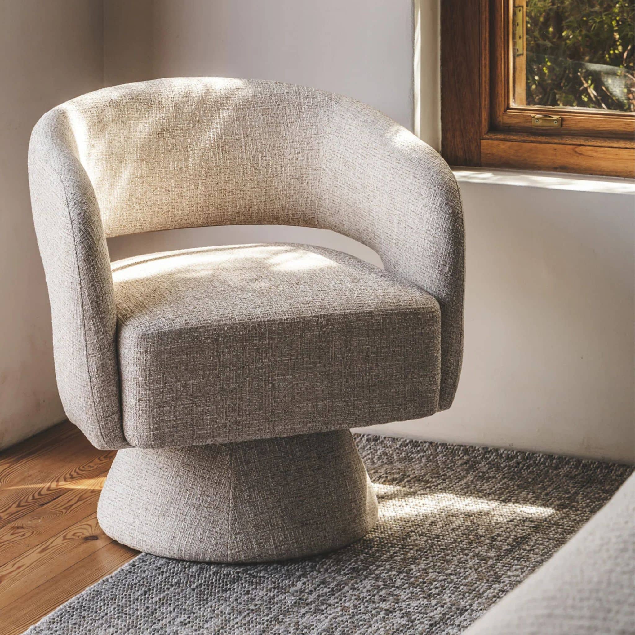 A modern barrel-style swivel armchair upholstered in textured light beige fabric, in a bedroom.