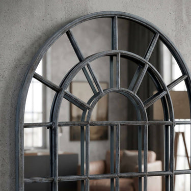 close up of Industrial Fenestra arched full-length mirror with a distressed black metal frame and sunburst windowpane grid, leaning against a textured grey concrete wall in a modern living room.