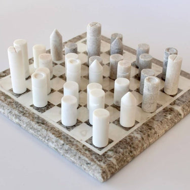 A minimalist geometric marble chess set from Woodka Interiors, cylindrical stone pieces in white and beige grey arranged on a square natural marble board.