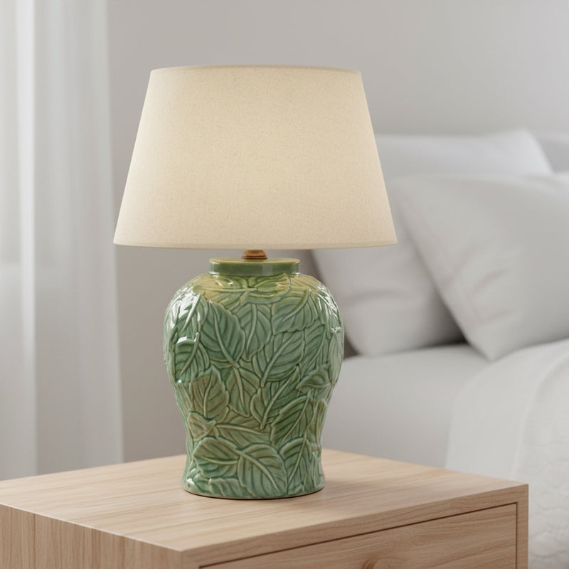 Green ceramic lamp with beige shade on a wooden bed side table 