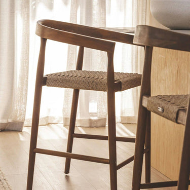 Two solid teak and handwoven rope counter chairs positioned next to a light oak wood kitchen island.