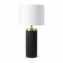 A modern table lamp with a black ribbed cylindrical base with a brushed gold neck collar and a white drum shade. 