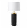 A modern table lamp with a black ribbed cylindrical base with a brushed gold neck collar and a white drum shade. 
