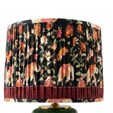 A pleated drum lampshade featuring a black background with a colorful print of elephants, flowers, and foliage, finished with a dark red ruffled band at the bottom and a gold-toned rim, shown on a white background.
