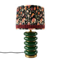 A table lamp with a glossy dark green stacked ceramic base and a pleated drum shade featuring a multicolor floral and animal print on a black background with a burgundy ruffled band.