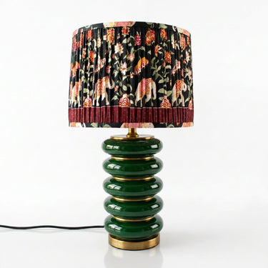 A table lamp with a glossy dark green stacked ceramic base and a pleated drum shade featuring a black background with a multicolor floral and animal print, finished with gold-tone metal trim.