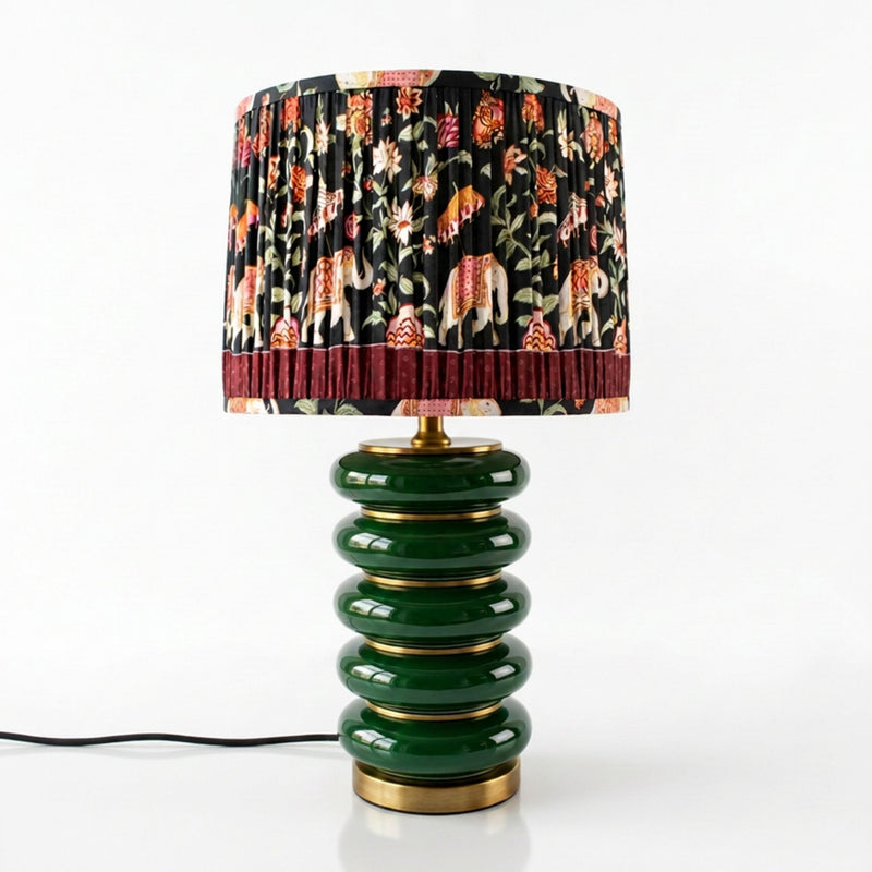 A table lamp with a glossy dark green stacked ceramic base and a pleated drum shade featuring a black background with a multicolor floral and animal print, finished with gold-tone metal trim.