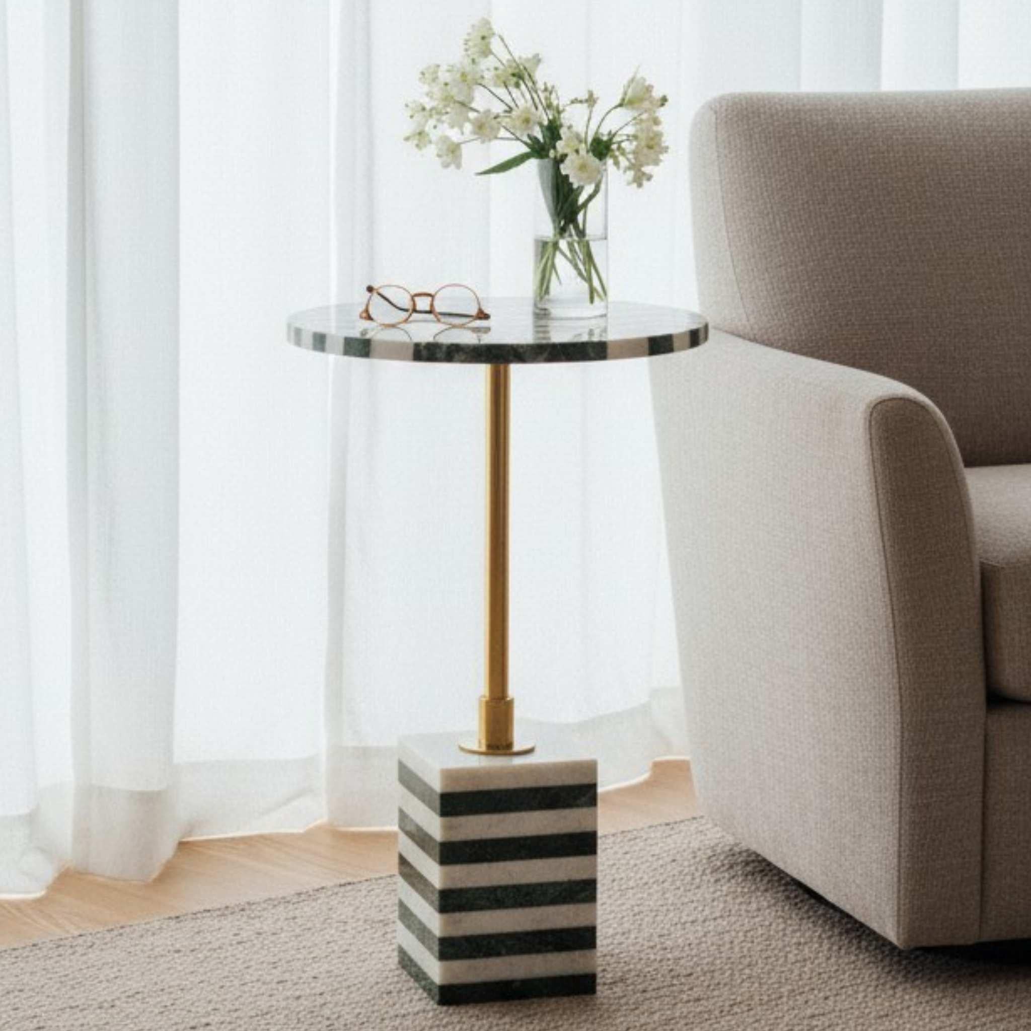 Small round table with marble top and striped base next to a beige armchair in a room with white curtains.