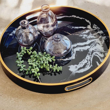Round black tray with gold rim featuring three glass bottles and greenery on a beige coffee table.