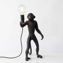 A black monkey figurine lamp holding a clear glass bulb, shown on a plain white background with the light turned on.