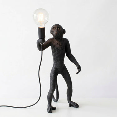 A black monkey figurine lamp holding a clear glass bulb, shown on a plain white background with the light turned on.