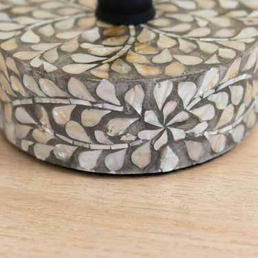 A close-up of a decorative round base  or the capiz petite accent table featuring a mother-of-pearl inlay pattern in beige, white, and gray tones, resting on a light wood surface.