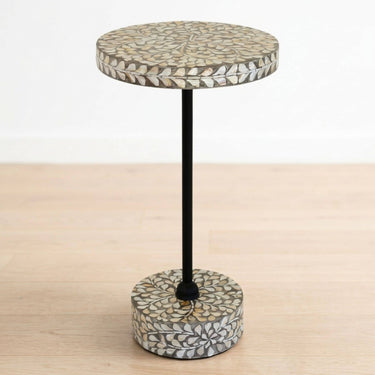 A small round side table with a black metal stand and a circular top and base covered in a mosaic inlay pattern of beige, brown, and white leaf-like shapes, shown on a light wood floor against a white wall.