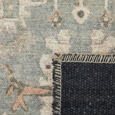 Close-up of a distressed area rug showing its faded beige and blue pattern, and a dark charcoal woven backing.
