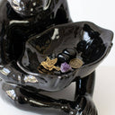A glossy black ceramic figurine of a seated ceramic monkey lamp holding a shallow dish with small stones, a gold bird charm, and a purple crystal.