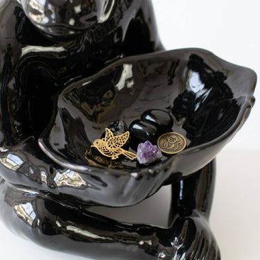 A glossy black ceramic figurine of a seated ceramic monkey lamp holding a shallow dish with small stones, a gold bird charm, and a purple crystal.