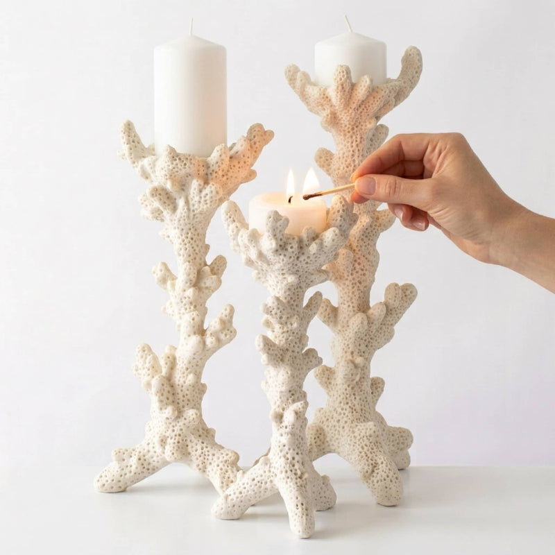 Three coral-shaped candle holders in a warm off-white tone, with two holding white taper candles and a hand lighting one with a match against a clean white background.