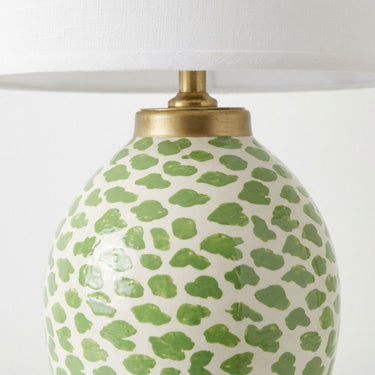 Close-up of a table lamp base with a glossy white ceramic body covered in irregular green spots, topped with a brass neck and a white fabric lampshade.