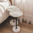 Two round marble-top side tables with black metal pedestal legs and round marble bases, placed beside a bed in a softly lit bedroom with curtains and wood flooring.