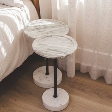 Two round marble-top side tables with black metal pedestal legs and round marble bases, placed beside a bed in a softly lit bedroom with curtains and wood flooring.