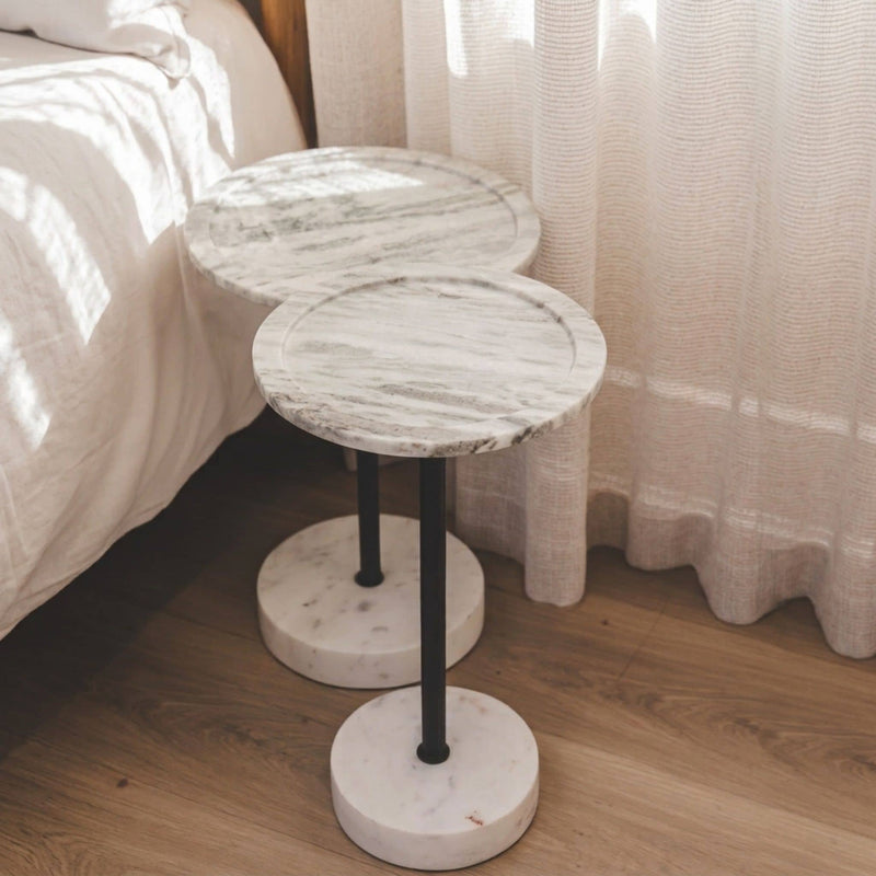 Two round marble-top side tables with black metal pedestal legs and round marble bases, placed beside a bed in a softly lit bedroom with curtains and wood flooring.