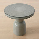 A round ceramic side table with a thick glazed top and a cylindrical pedestal base, shown on a light wood surface.