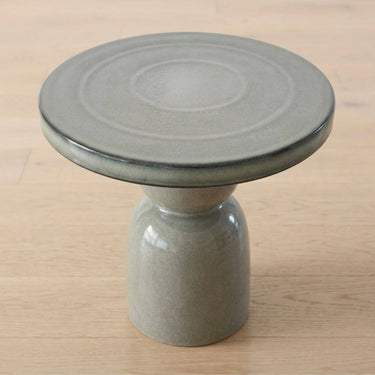 A round ceramic side table with a thick glazed top and a cylindrical pedestal base, shown on a light wood surface.