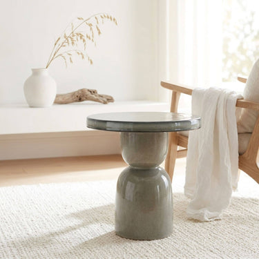 A round grey ceramic side table with a sculptural pedestal base, styled in a bright, minimalist room next to a wooden chair with a white throw blanket.