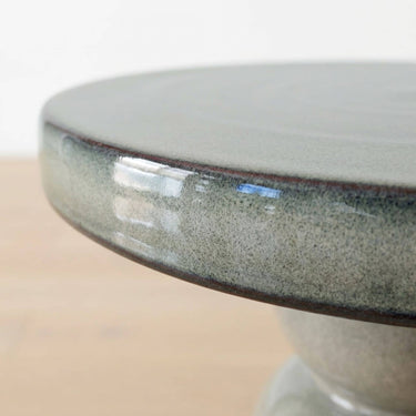 Close-up of a round ceramic pedestal side table round top edge  with a speckled grey glaze and a dark rim, photographed indoors against a light neutral background.