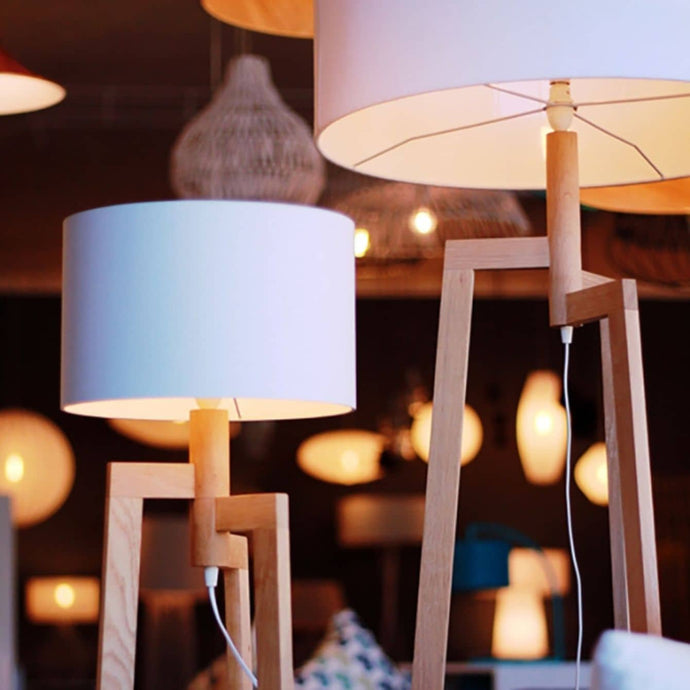 Two modern floor lamps with white cylindrical shades and wooden tripod bases are displayed in a room with several other illuminated lamps and decorative light fixtures in the background.