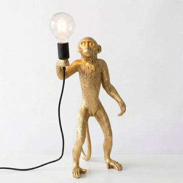 A gold-finished monkey figurine holding an exposed light bulb, with a black cord and socket, photographed against a plain light background.