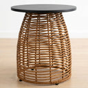 A round side table with a dark charcoal black top and a natural brown woven rattan base, shown on a light wood floor against a white wall.