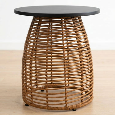 A round side table with a dark charcoal black top and a natural brown woven rattan base, shown on a light wood floor against a white wall.