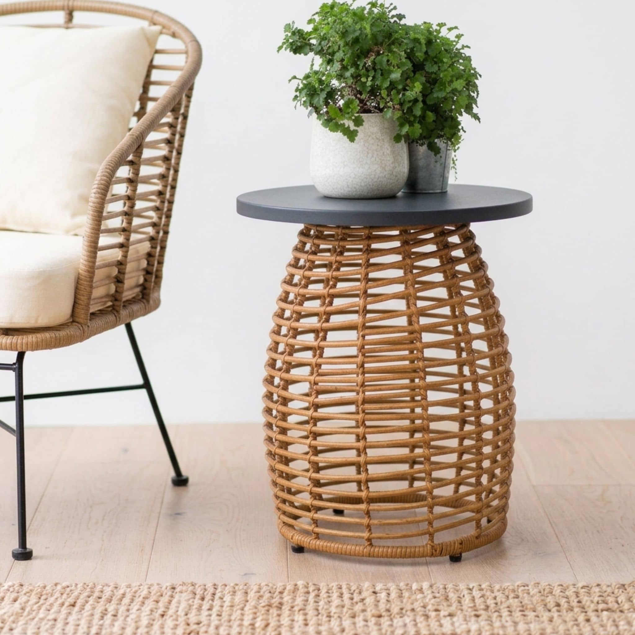 A round side table with a dark gray top and a natural woven rattan basket base, styled with a potted green plant. A matching woven armchair with a cream cushion is partially visible on the left against a clean white wall and light wood floor.
