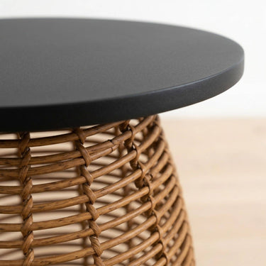 Close-up of a round black side table with a woven natural brown wicker base, shown against a light neutral background.