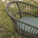 Close-up of a woven outdoor chair with a curved back and seat cushion, shown on grass outdoors.