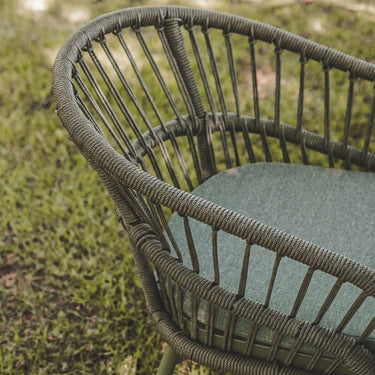 Close-up of a woven outdoor chair with a curved back and seat cushion, shown on grass outdoors.