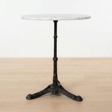 A round white marble bistro table with a black cast iron pedestal base, shown on a light wood floor against a white wall.