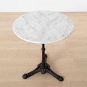A round white marble table with a black cast iron pedestal base, set on a light wood floor against a white wall.