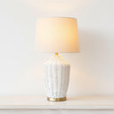 Illuminated white rattan bedside lamp with a brass accent providing a warm ambient glow in a bright, airy room.