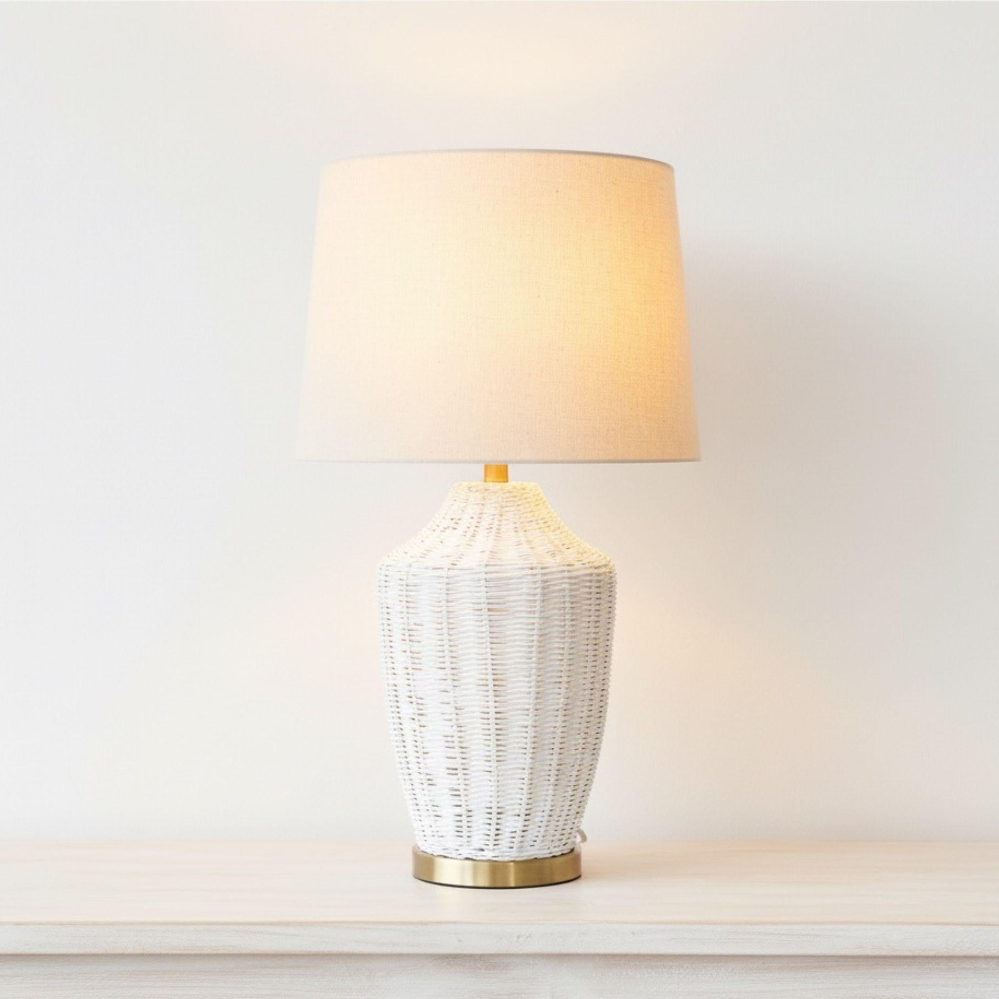 Illuminated white rattan bedside lamp with a brass accent providing a warm ambient glow in a bright, airy room.