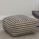 Striped wool and jute pouf ottoman placed next to a brown chair on a textured rug.