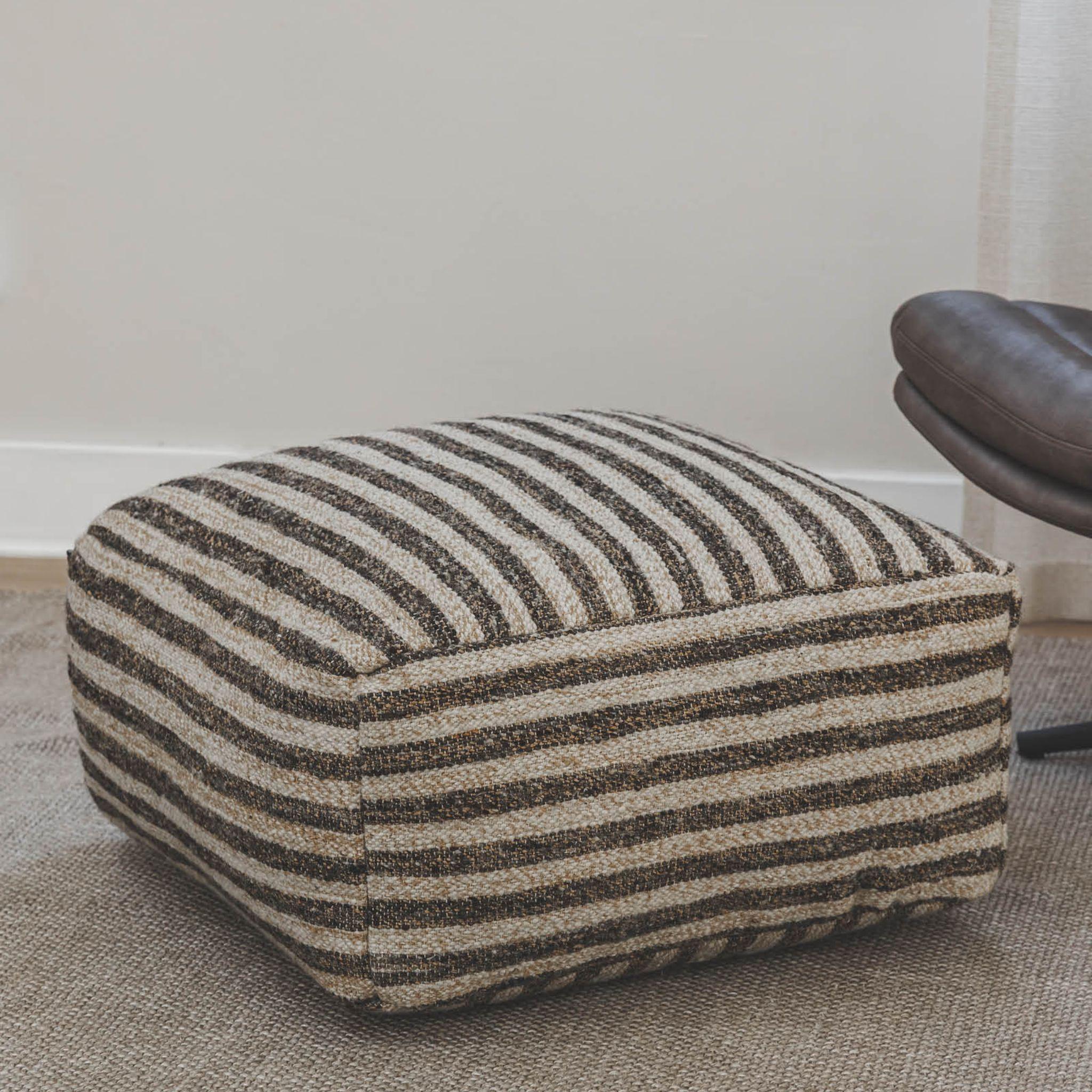 Striped wool and jute pouf ottoman placed next to a brown chair on a textured rug.