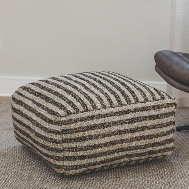 Striped wool and jute pouf ottoman placed next to a brown chair on a textured rug.