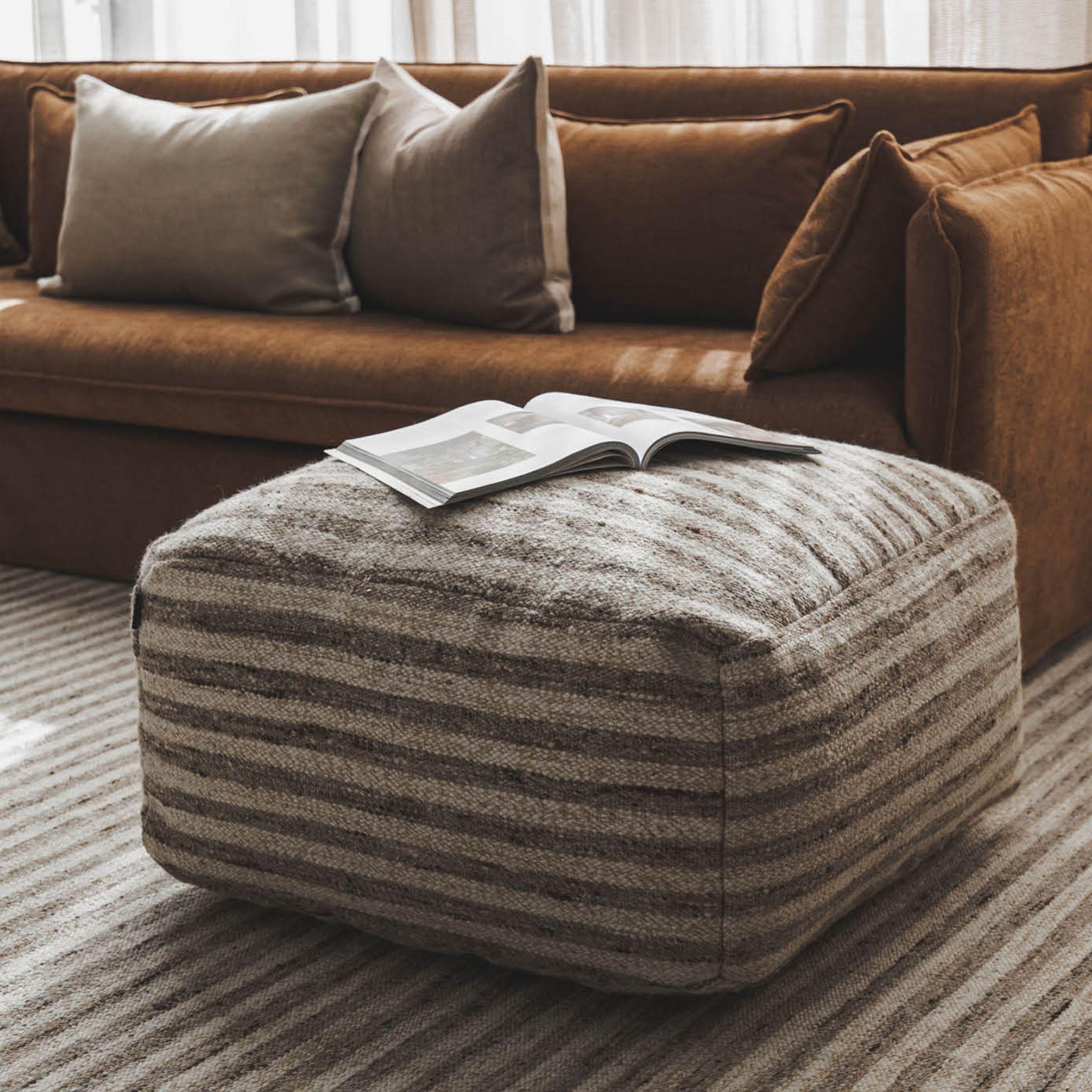 Paralla striped wool-jute square pouf ottoman on a matching rug in front of a brown sofa.