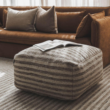 Paralla striped wool-jute square pouf ottoman on a matching rug in front of a brown sofa.