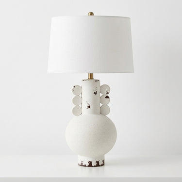 distressed table lamp with a textured  ceramic base, a white drum shade, and brass-toned metal accents.
