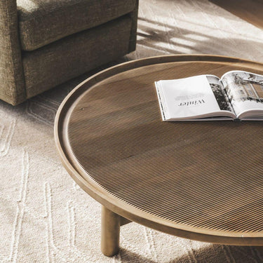 Sandpiper wood coffee table with a mango wood round top in a vintage finish  styled in modern living room with chair and beige rug.