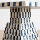 A close-up of a table base with a geometric checkered pattern made of alternating dark navy and  white capiz inlays, set against a bright white background.