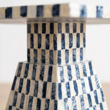 A close-up of a table base with a geometric checkered pattern made of alternating dark navy and  white capiz inlays, set against a bright white background.